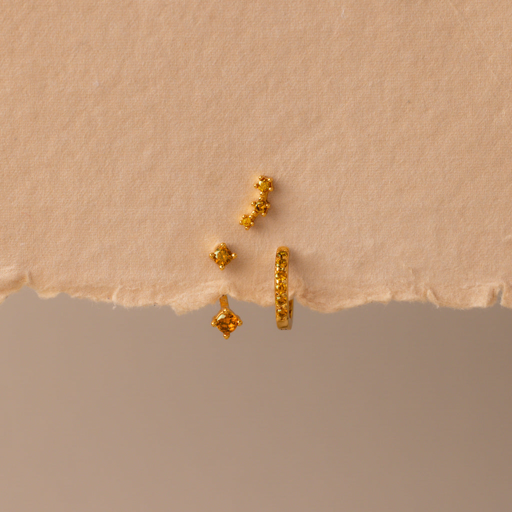 The Topaz Earring Trio Set features four small gold earrings with yellow gemstones, beautifully displayed on a torn piece of beige textured paper—perfect as a birthstone earring set.