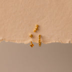 The Topaz Earring Trio Set features four small gold earrings with yellow gemstones, beautifully displayed on a torn piece of beige textured paper—perfect as a birthstone earring set.