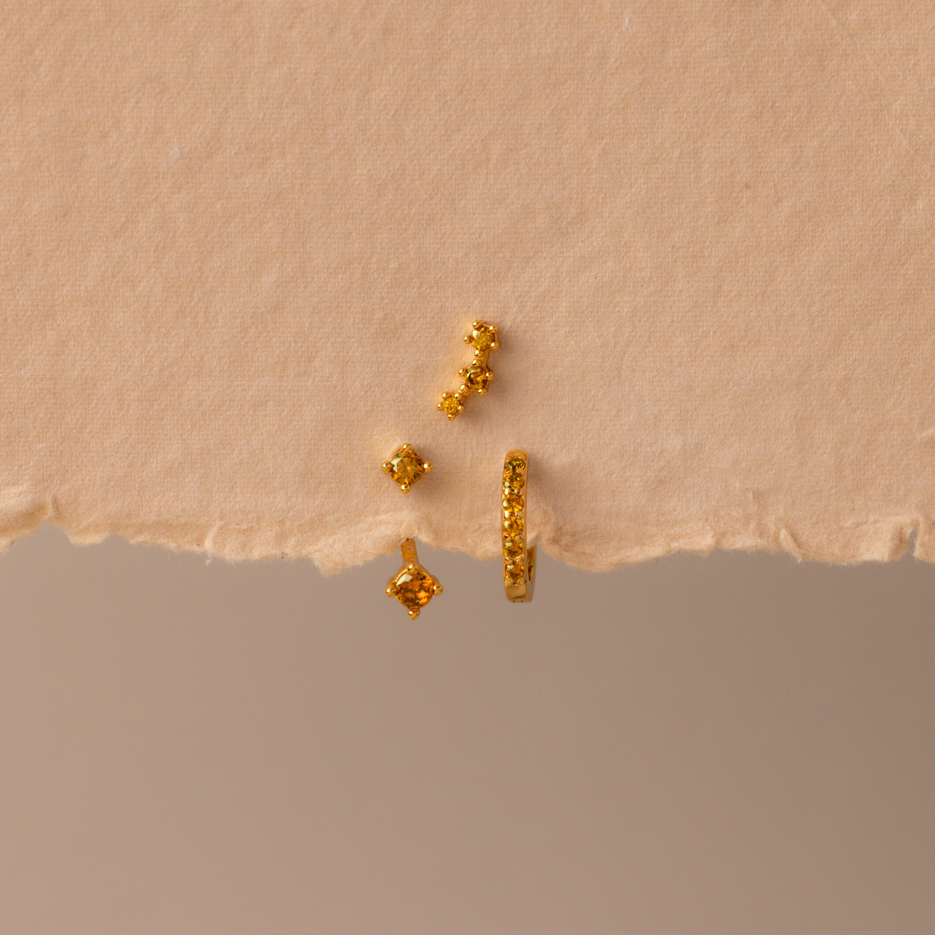 The Topaz Earring Trio Set features four small gold earrings with yellow gemstones, beautifully displayed on a torn piece of beige textured paper—perfect as a birthstone earring set.