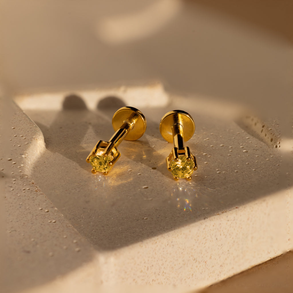 A pair of Topaz Flatback Studs featuring yellow gemstones is displayed on a textured beige surface.