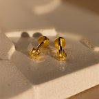 A pair of Topaz Flatback Studs featuring yellow gemstones is displayed on a textured beige surface.