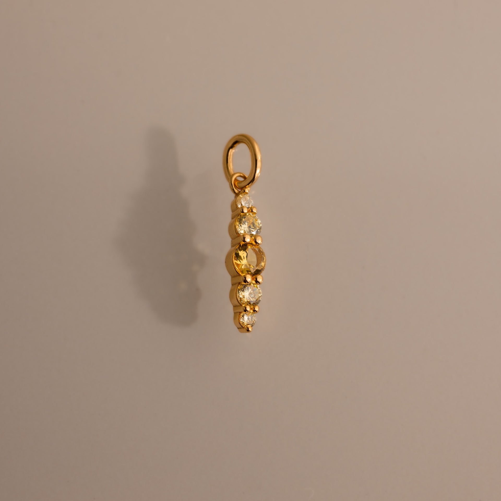 Ombre Lace Topaz Charm: a gold pendant featuring four white gemstones set vertically, displayed on a plain light brown background—an elegant birthstone accessory.
