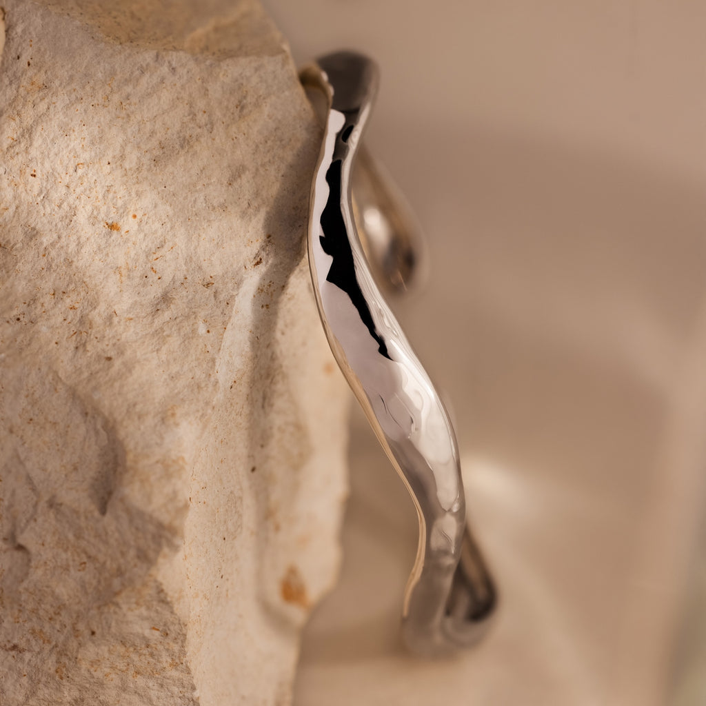 The Organic Wavy Cuff Bracelet in Silver, featuring sculpted curves, rests against a textured, light-colored stone surface.