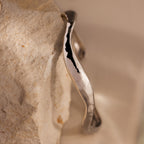 The Organic Wavy Cuff Bracelet in Silver, featuring sculpted curves, rests against a textured, light-colored stone surface.