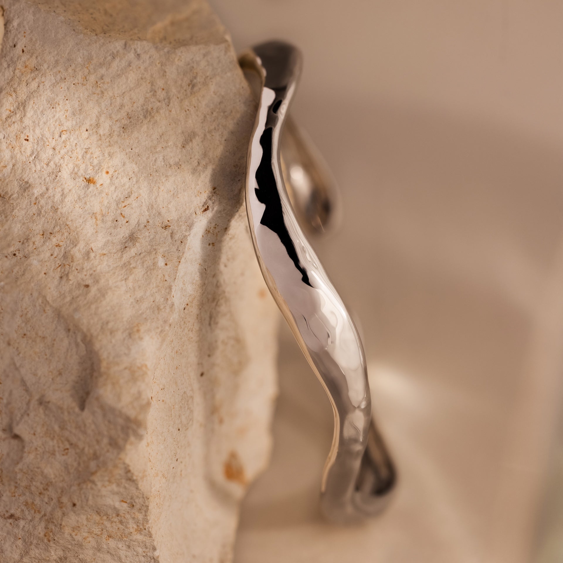 The Organic Wavy Cuff Bracelet in Silver, featuring sculpted curves, rests against a textured, light-colored stone surface.