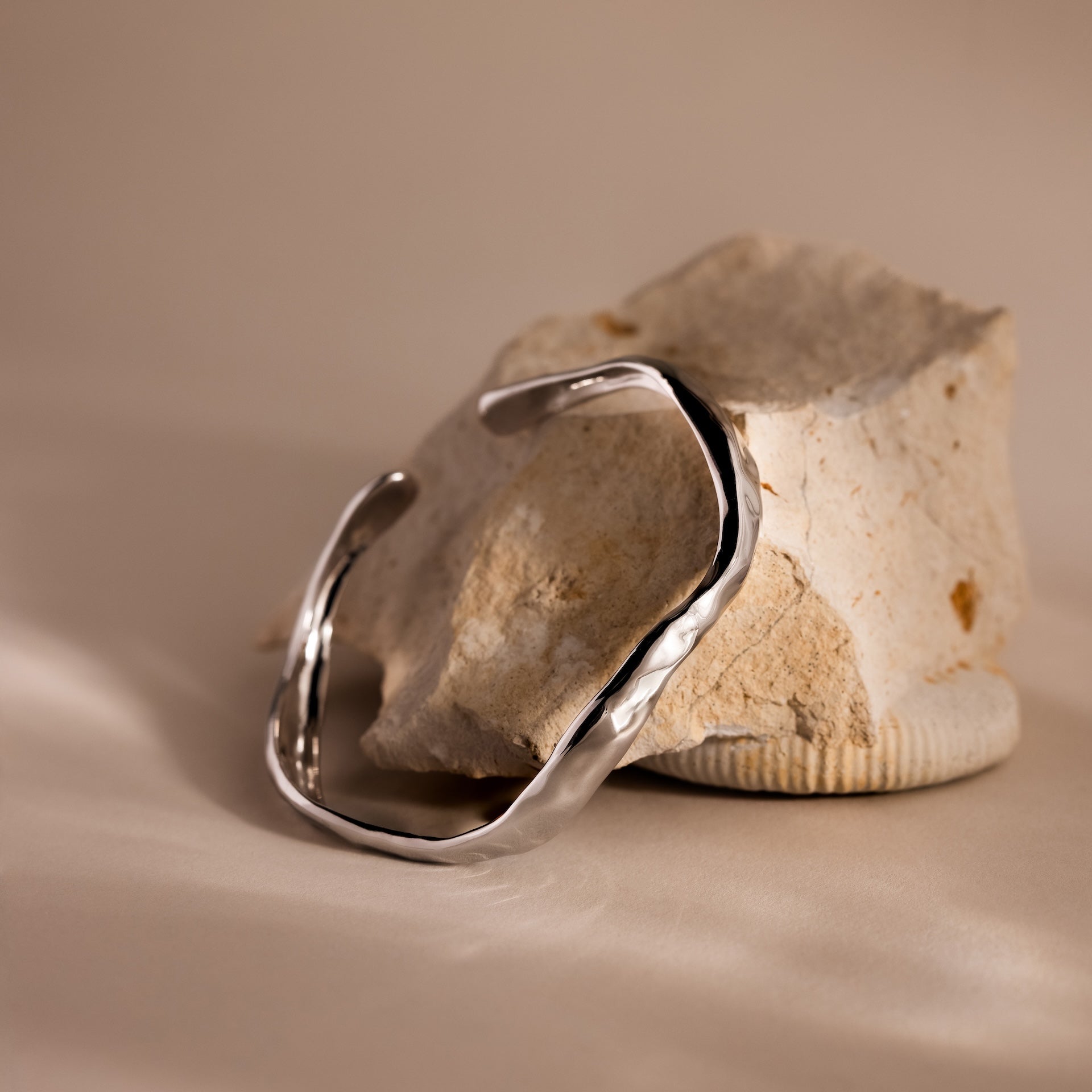 The Organic Wavy Cuff Bracelet in silver with a textured finish is displayed on a beige stone, set against a soft, neutral background.