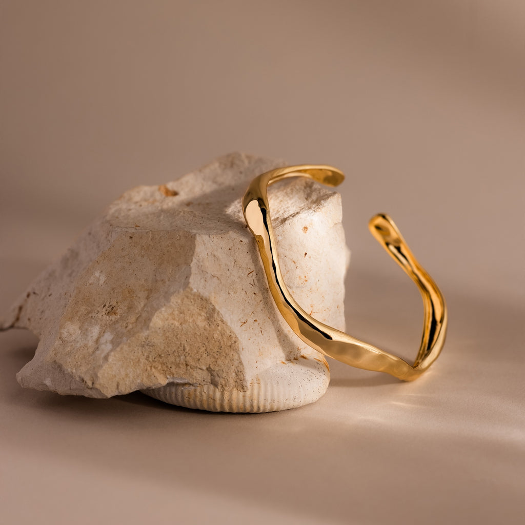 The Organic Wavy Cuff Bracelet in 18K Gold, featuring sculpted curves, rests elegantly on a textured, light rock against a neutral backdrop—an effortlessly timeless accessory.