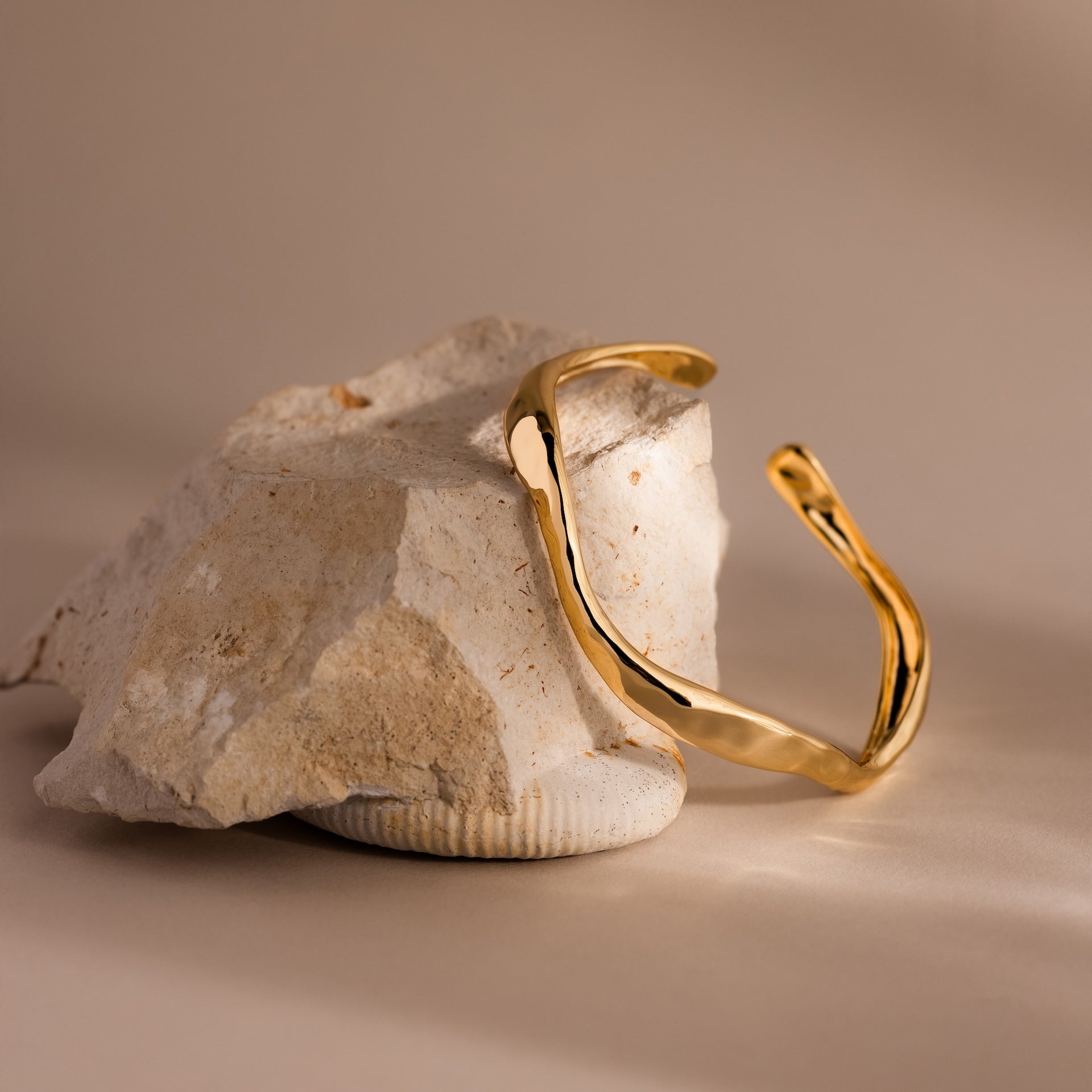 The Organic Wavy Cuff Bracelet in 18K Gold, featuring sculpted curves, rests elegantly on a textured, light rock against a neutral backdrop—an effortlessly timeless accessory.