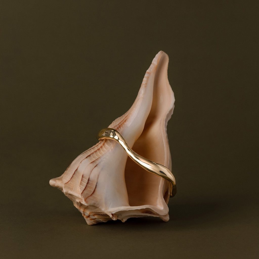 The Organic Wavy Cuff Bracelet, featuring sculpted gold curves, is elegantly displayed draped over a pink seashell on a plain brown background.