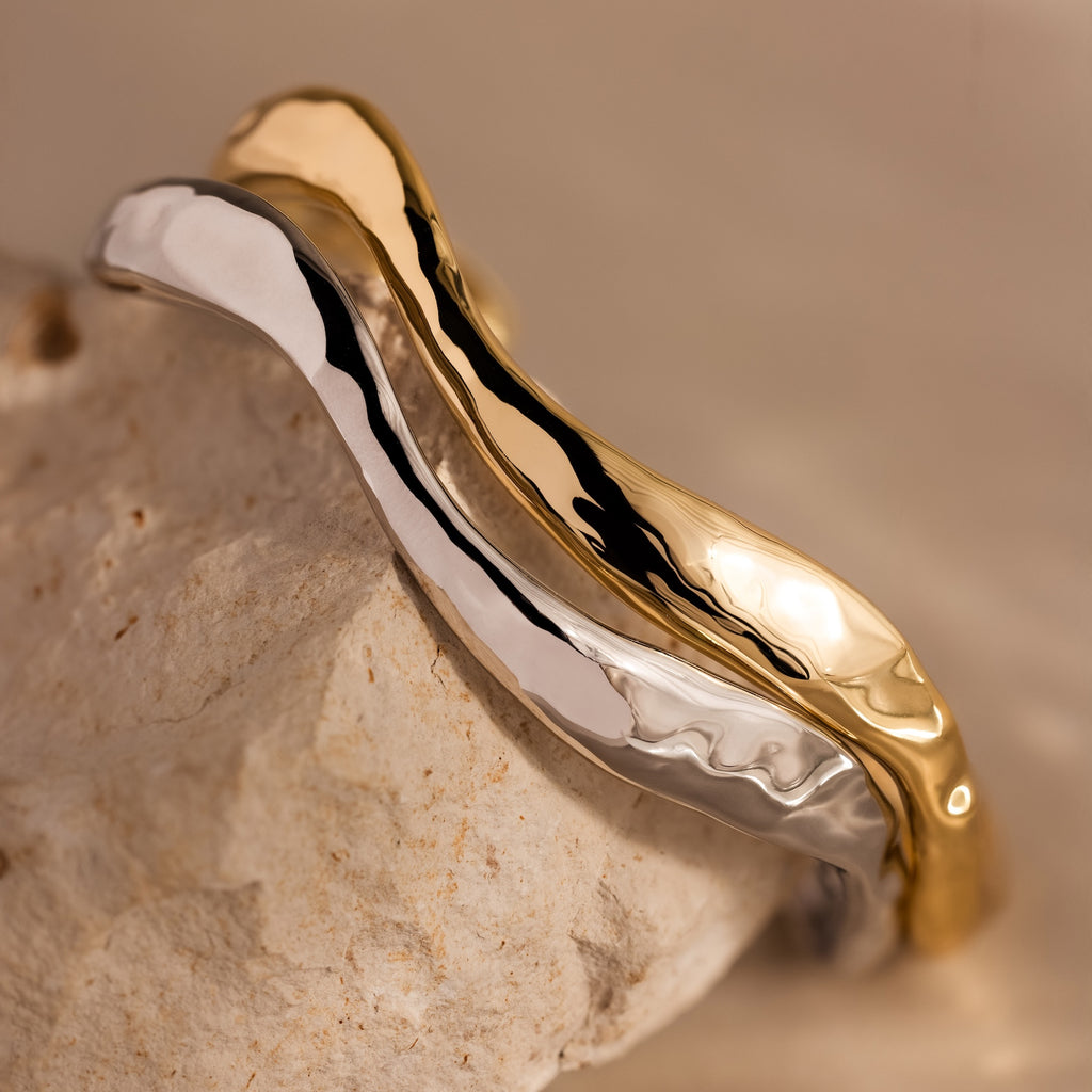 The Organic Wavy Cuff Bracelets, one in gold and one in silver, feature sculpted curves and rest elegantly on a textured beige stone.