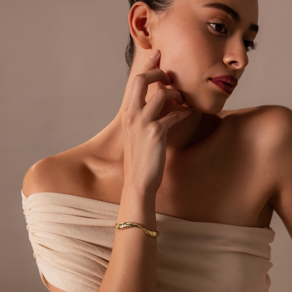 A woman in an off-shoulder beige top touches her face, wearing the Organic Wavy Cuff Bracelet in 18K Gold—a timeless piece highlighting her sculpted curves as she looks to the side.
