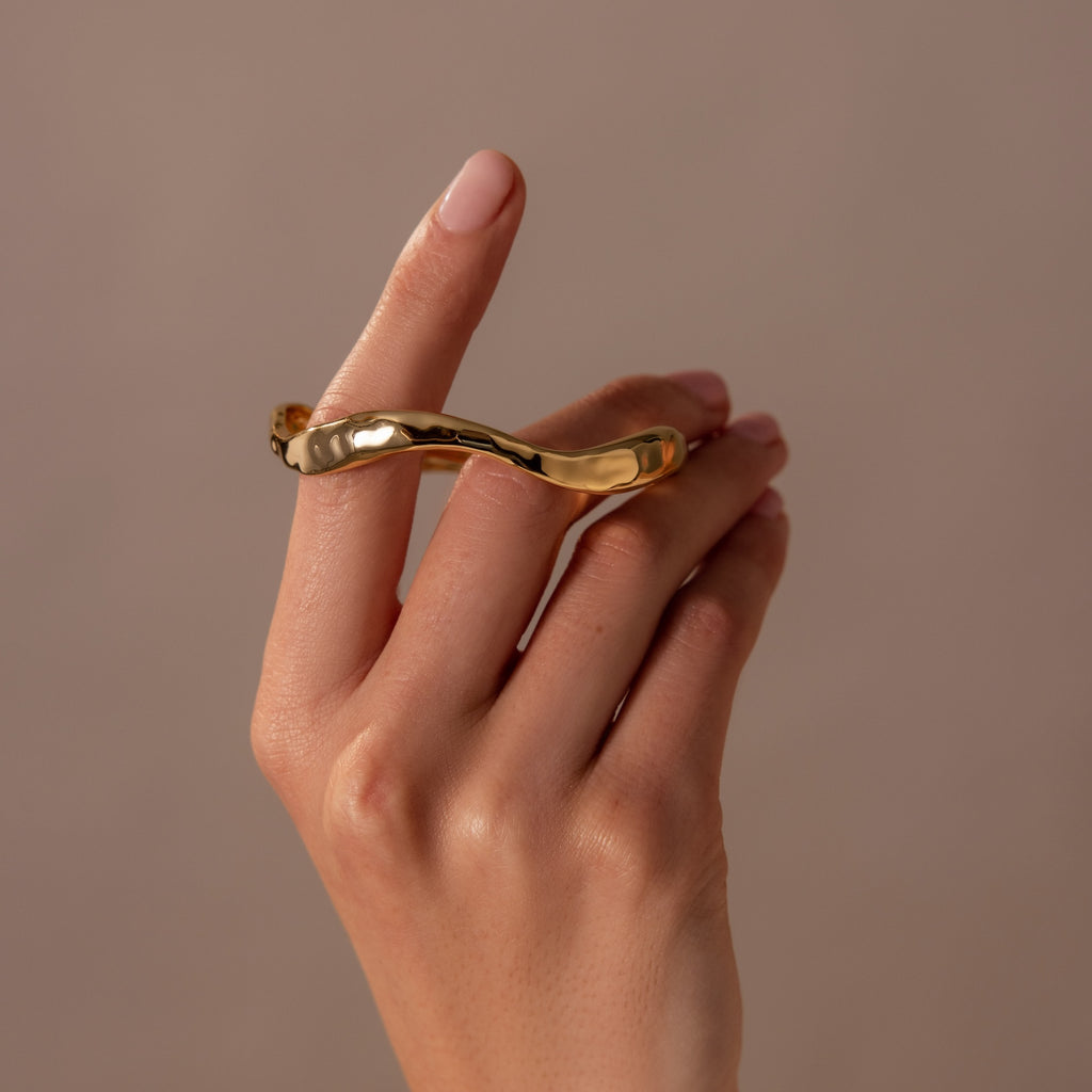 An arm with nude nails showcases the Organic Wavy Cuff Bracelet in 18K Gold against a beige background—a sculptural, timeless statement piece.