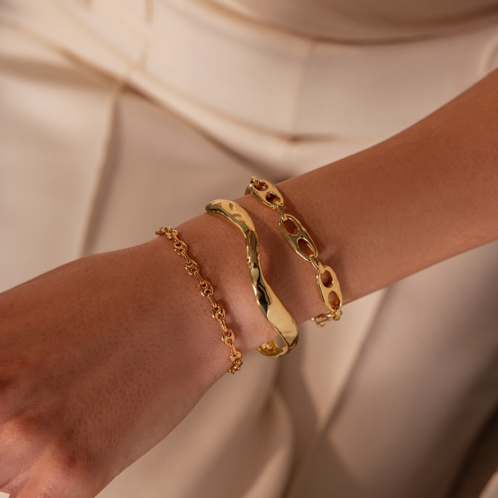 A hand adorned with three gold bracelets, including the Organic Wavy Cuff Bracelet in 18K Gold with its sculpted curves and unique textures, styled with chain designs against a beige outfit—a truly timeless accessory.