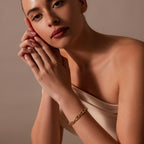 A woman in an off-shoulder top poses with hands clasped, highlighting the Organic Mariner Chain Bracelet in 18K Gold—a sculptural essential—against a neutral background.