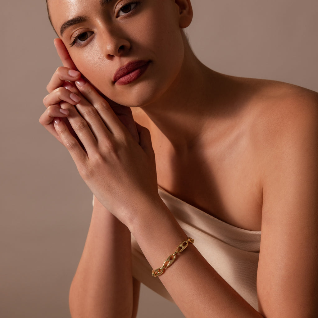 A woman in an off-shoulder top wears the Organic Mariner Chain Bracelet, resting her head on her clasped hands—a stylish touch of contemporary jewelry.