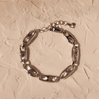 The Organic Mariner Chain Bracelet in Silver, featuring oval links, is displayed on a textured beige surface, offering a timeless style perfect for any occasion.