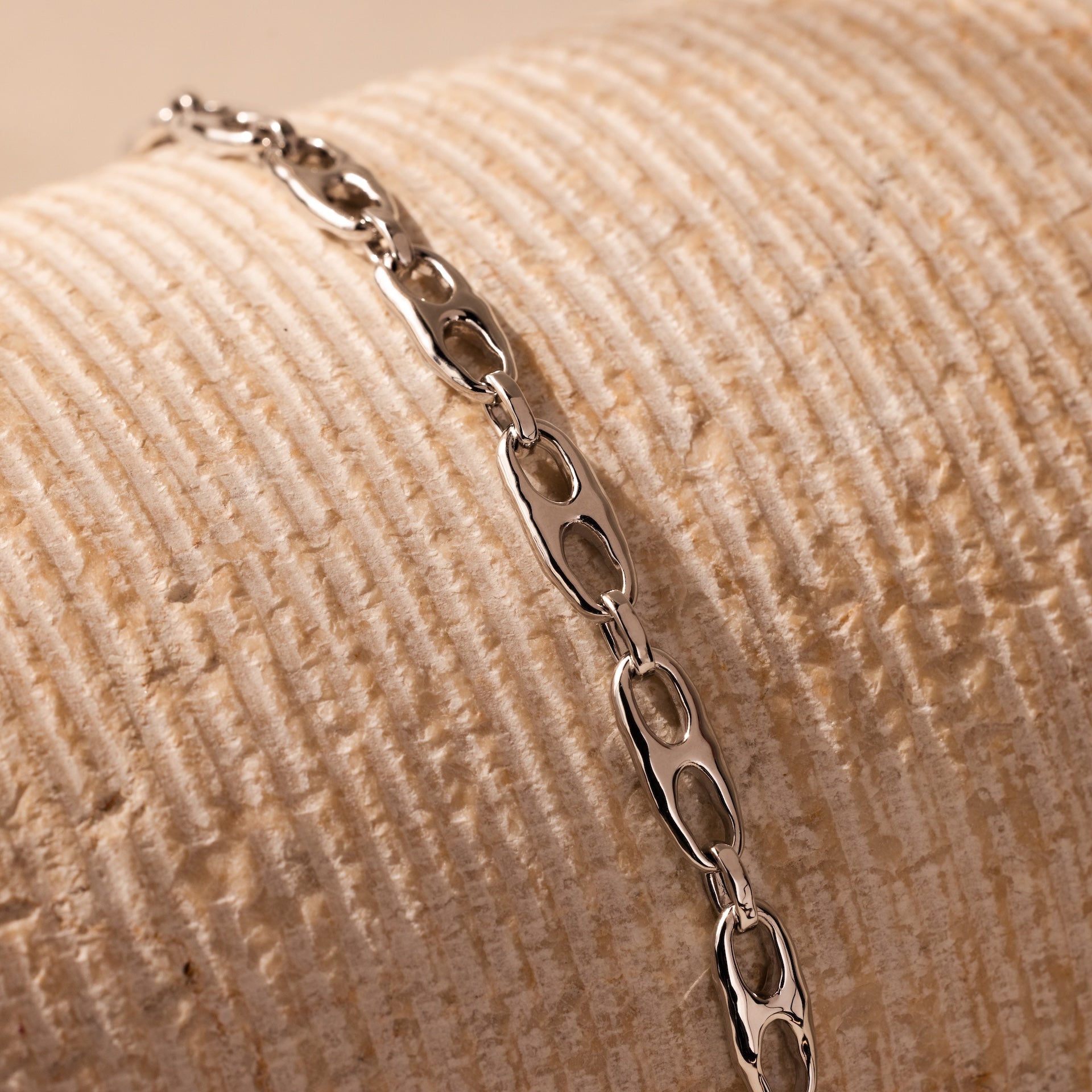 The Organic Mariner Chain Bracelet in Silver features sculptural oval links, elegantly displayed on a textured beige surface.