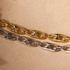 The Organic Mariner Chain Bracelet in 18K Gold and a silver sculptural chain bracelet are displayed on a beige textured surface, highlighting modern jewelry design.