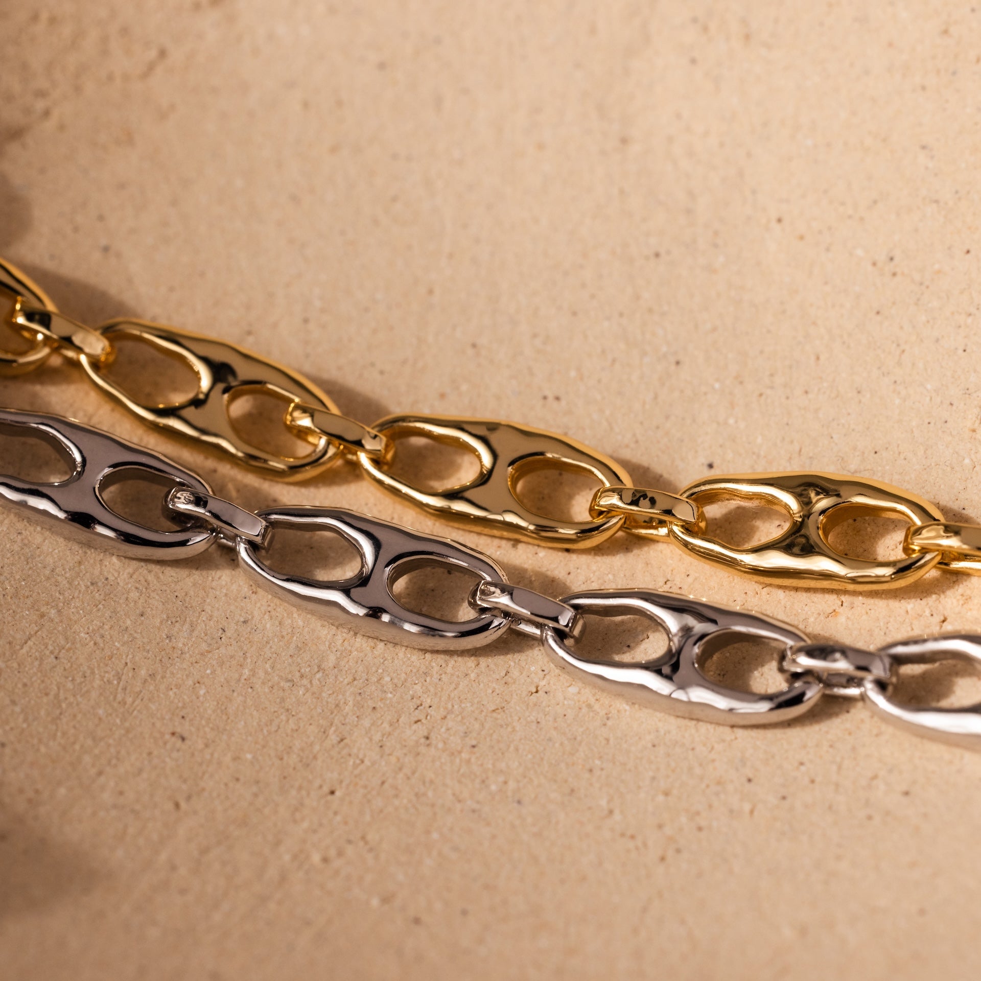 The Organic Mariner Chain Bracelet in 18K Gold and a silver sculptural chain bracelet are displayed on a beige textured surface, highlighting modern jewelry design.