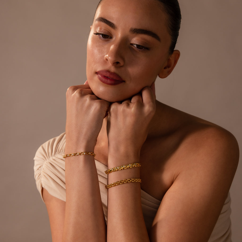 A woman with a neutral expression wears an off-shoulder beige top and showcases the Diamond Elephant Link Bracelet along with other gold bracelets on both wrists.
