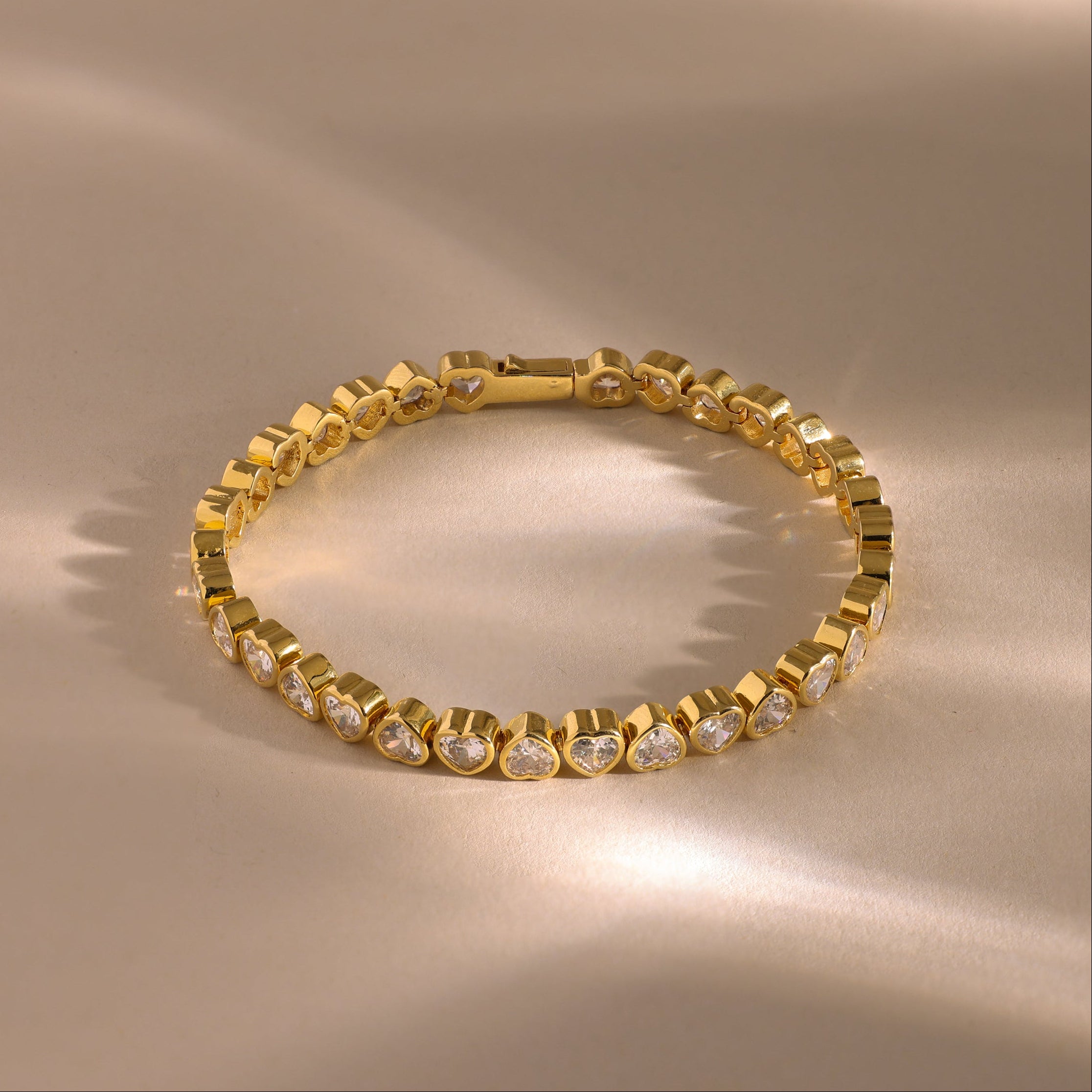 The Heart Tennis Bracelet in 18K Gold is displayed on a beige surface with soft, shimmering light—an exquisite piece of elegant jewelry perfect for any occasion.