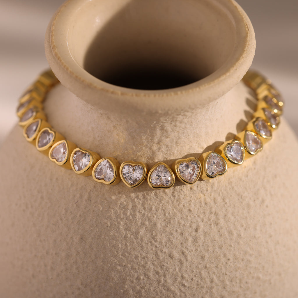 The Heart Tennis Bracelet in 18K Gold, adorned with elegant heart-shaped stones, is draped around the neck of a textured beige ceramic vase.