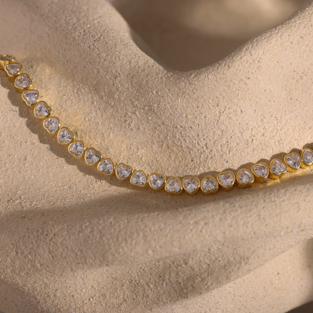 Elegant jewelry showcasing the Heart Tennis Bracelet in 18K Gold, adorned with heart-shaped diamonds and displayed on a textured beige stone surface.