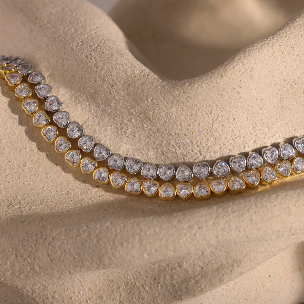 A gold and silver diamond necklace rests on a textured beige surface, elegantly paired with the Heart Tennis Bracelet—a sophisticated accessory featuring heart-shaped stones perfect for complementing any jewelry collection.