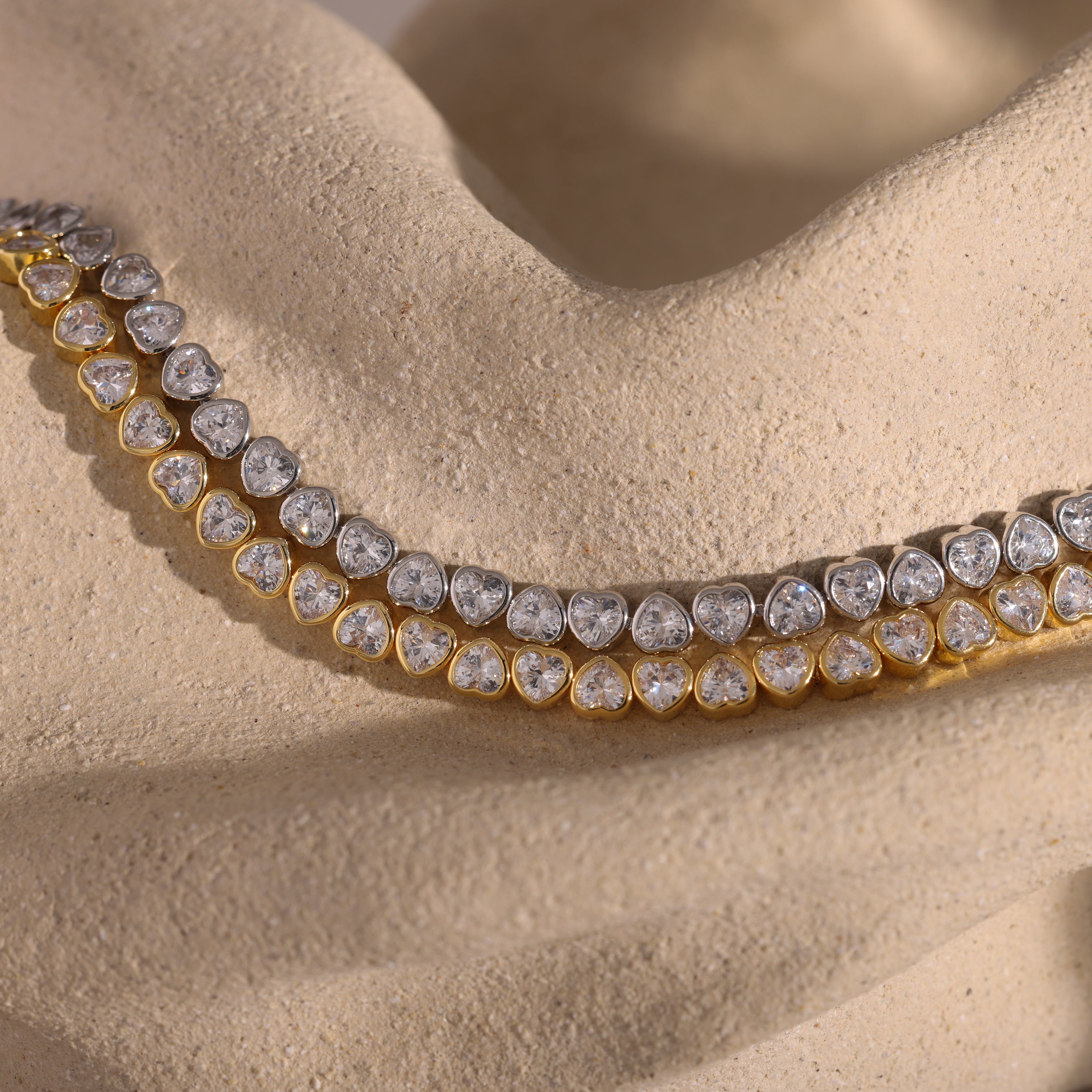 A gold and silver diamond necklace rests on a textured beige surface, elegantly paired with the Heart Tennis Bracelet—a sophisticated accessory featuring heart-shaped stones perfect for complementing any jewelry collection.