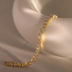 The Heart Tennis Bracelet, an elegant gold piece adorned with sparkling stones, is gracefully displayed on a smooth white ceramic surface.