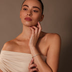 A woman in an off-shoulder beige dress touches her neck, highlighting the Heart Tennis Bracelet in 18K Gold against a neutral brown background.