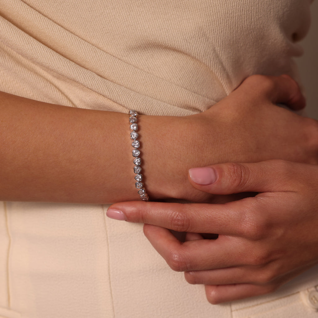 A woman dressed in a cream-colored outfit holds her arm, showcasing the Heart Tennis Bracelet in Silver with heart-shaped stones.