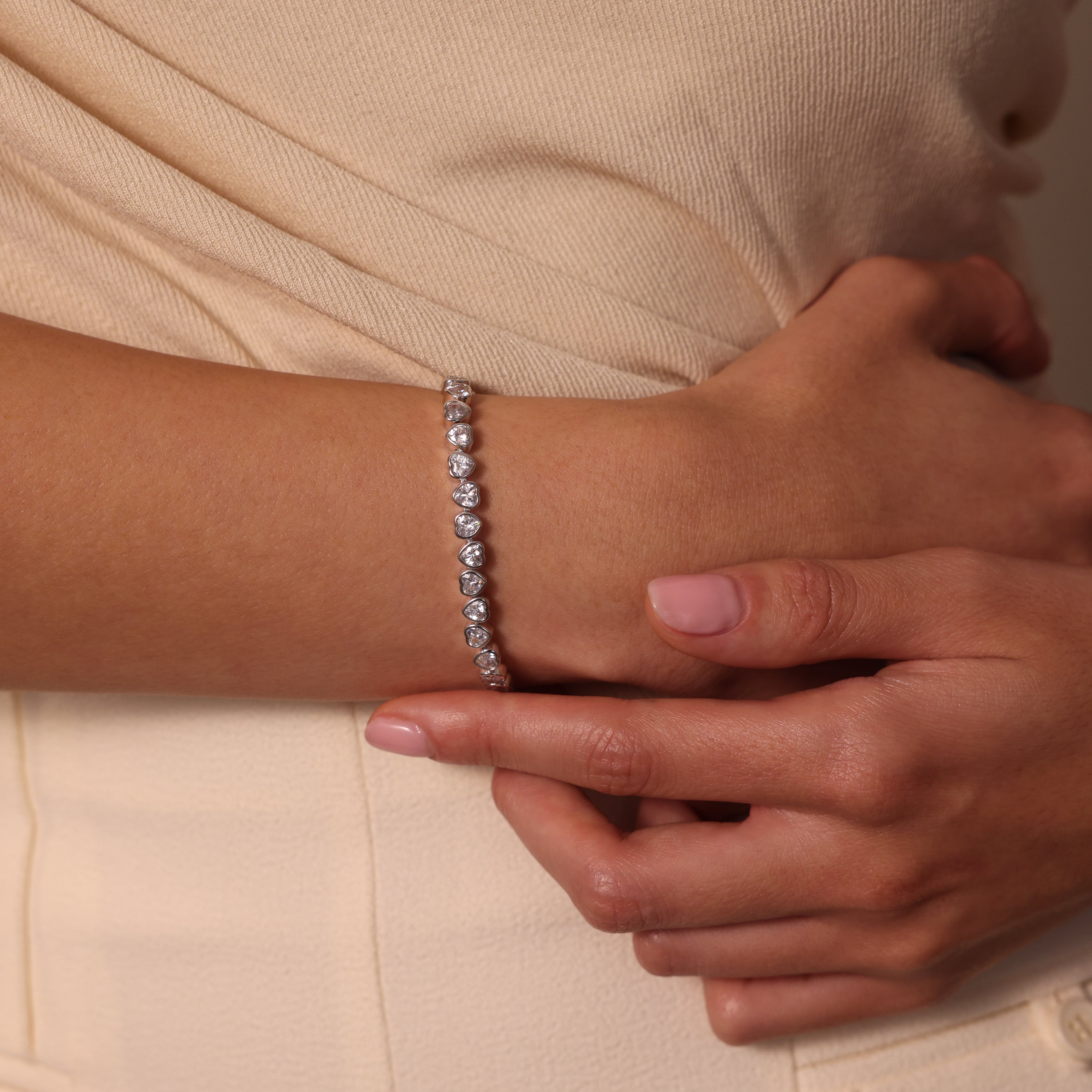 A woman dressed in a cream-colored outfit holds her arm, showcasing the Heart Tennis Bracelet in Silver with heart-shaped stones.