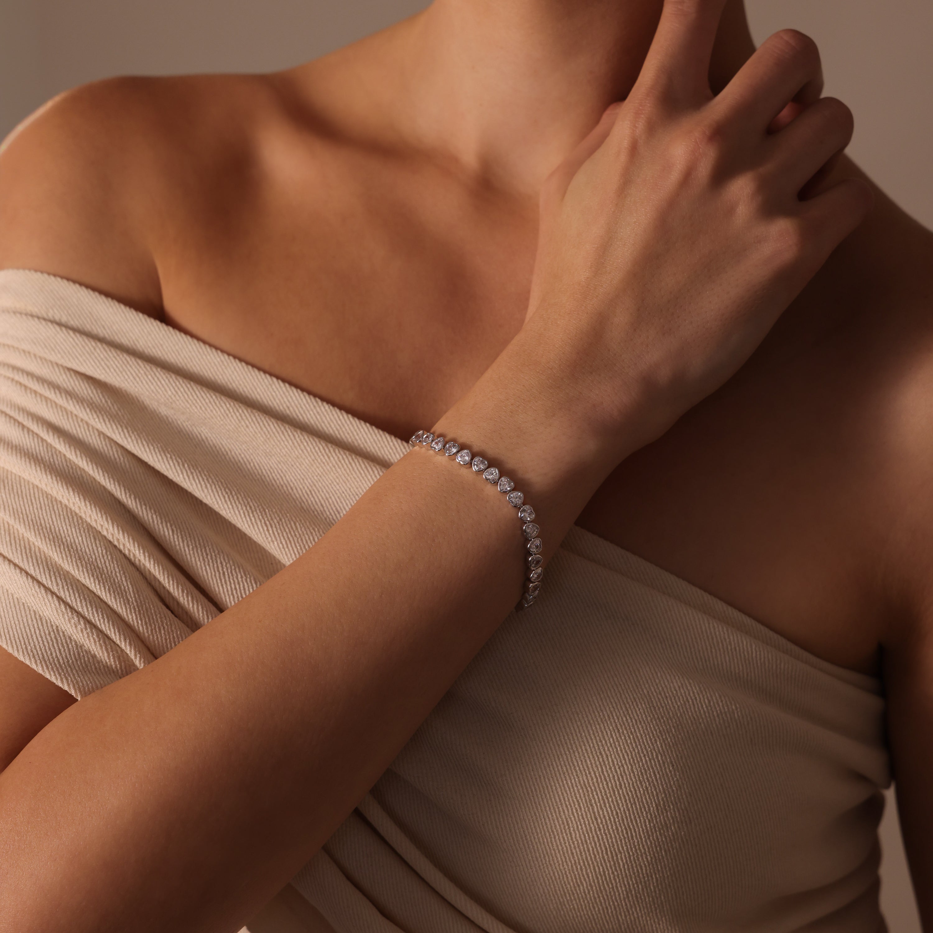 A woman in an off-shoulder beige dress radiates timeless beauty, accessorized with the Heart Tennis Bracelet in Silver adorned with heart-shaped stones.