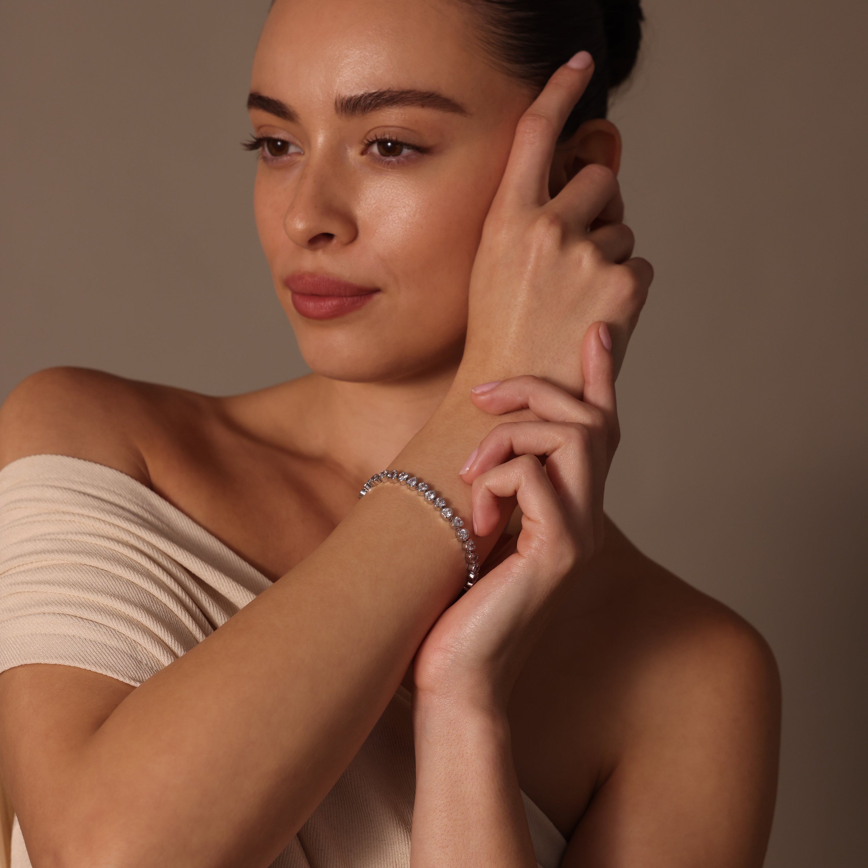 A woman in an off-shoulder top touches her face while wearing the Heart Tennis Bracelet in Silver, adorned with heart-shaped stones, and looks to the side with timeless elegance.