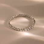 The Heart Tennis Bracelet in Silver features gemstone hearts, beautifully showcased on a beige surface with soft reflections that highlight its timeless charm.