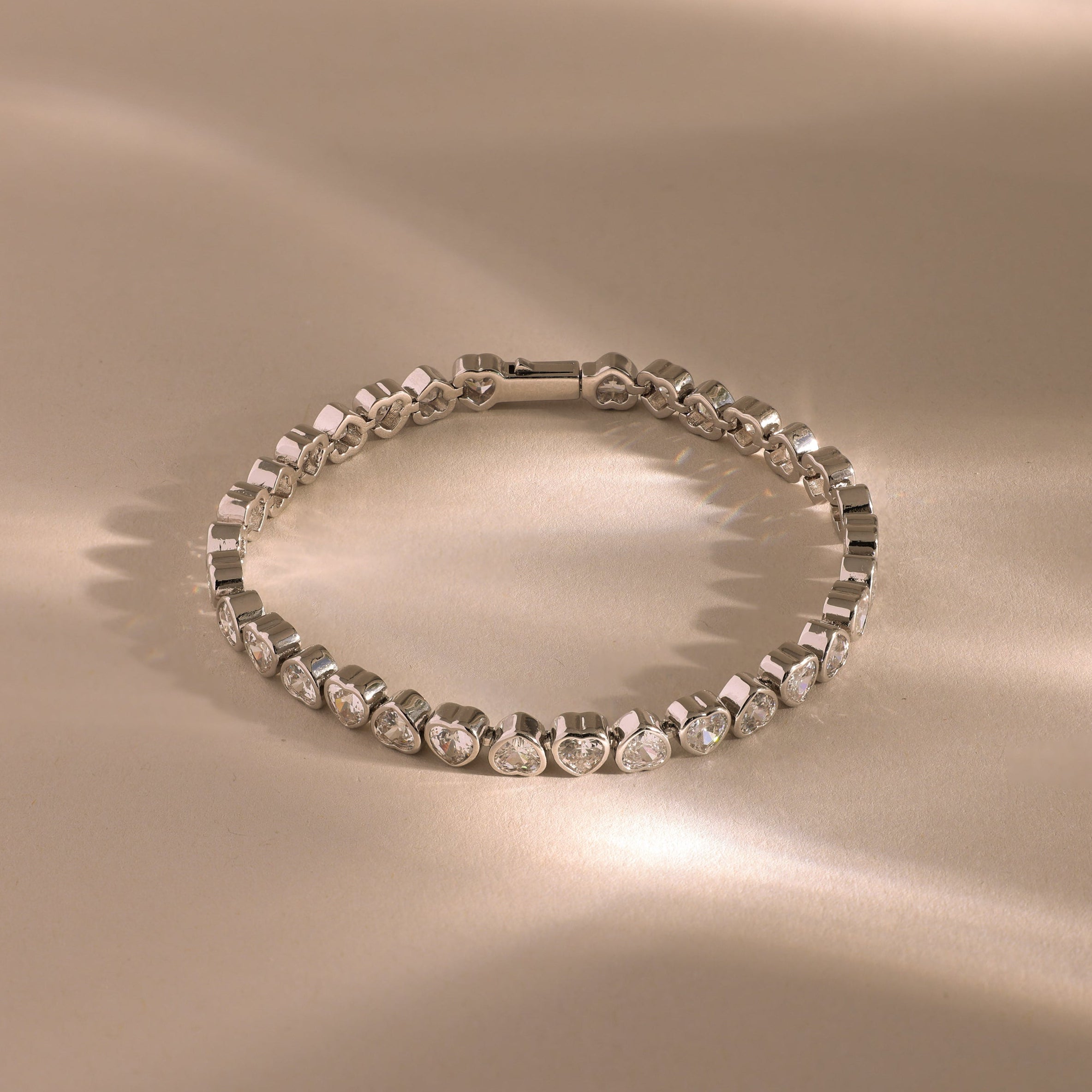 The Heart Tennis Bracelet in Silver features gemstone hearts, beautifully showcased on a beige surface with soft reflections that highlight its timeless charm.