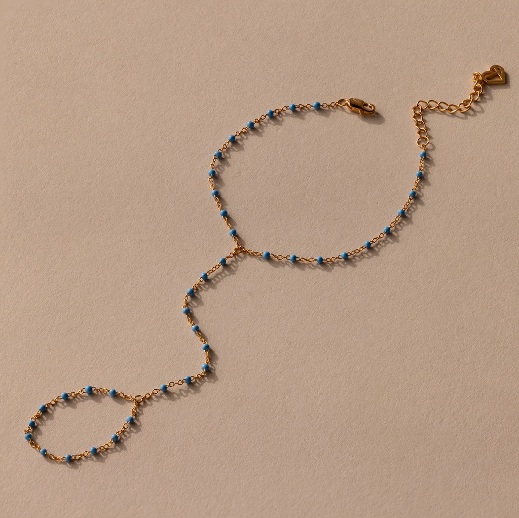 Ocean Beaded Hand Chain Bracelet in 18K Gold with blue beads, featuring a turquoise bracelet and ring linked by a delicate chain, showcased on a beige background.