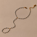 The Ocean Beaded Hand Chain Bracelet features a delicate gold chain with turquoise beads, an adjustable clasp, and ring detail, beautifully displayed on a neutral background.