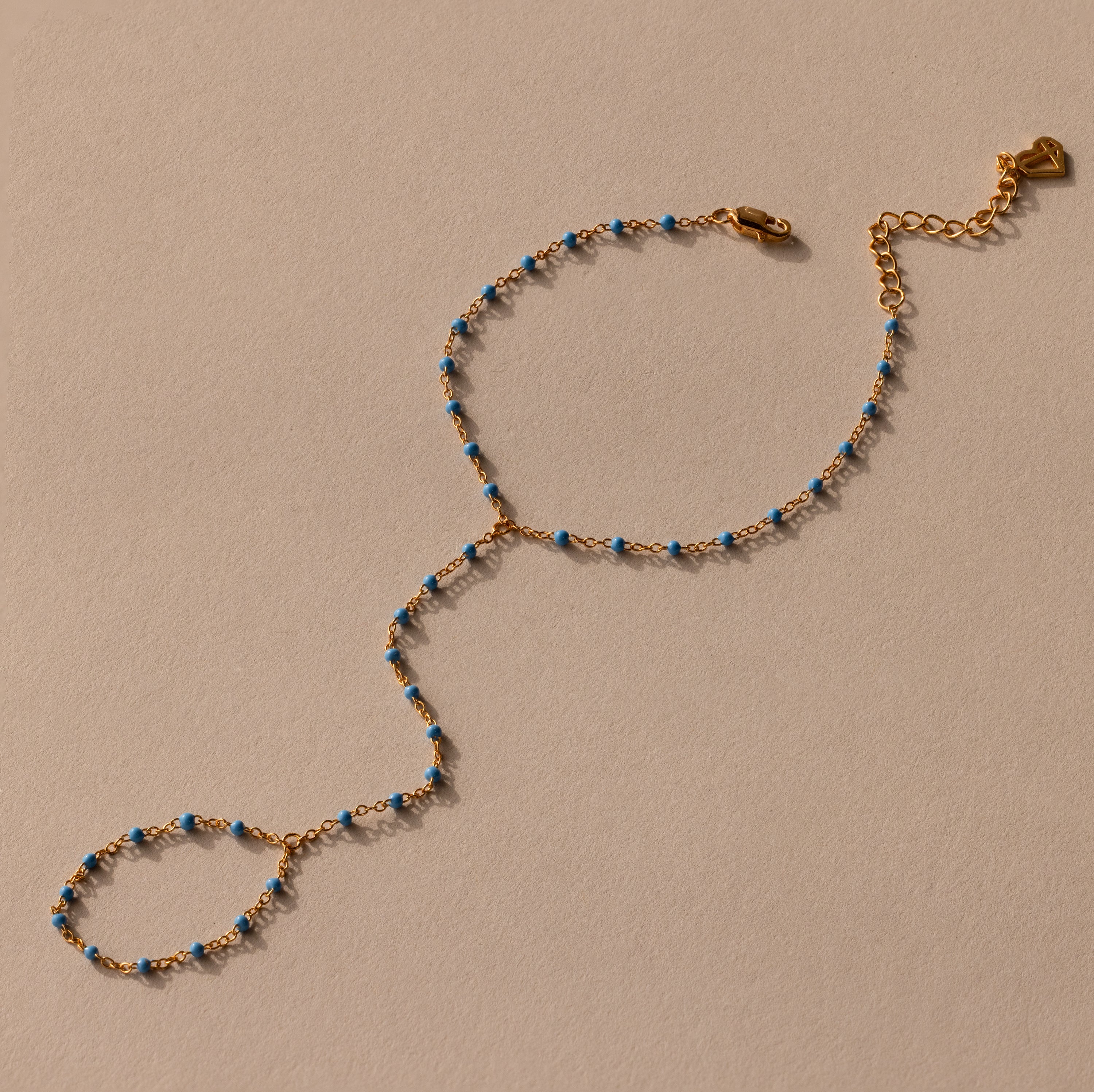 The Ocean Beaded Hand Chain Bracelet features a delicate gold chain with turquoise beads, an adjustable clasp, and ring detail, beautifully displayed on a neutral background.