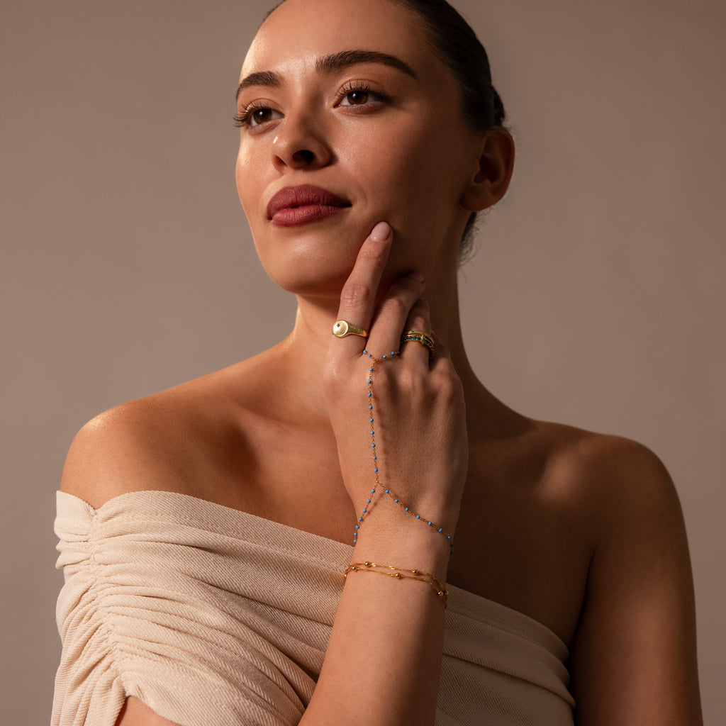 Woman in an off-shoulder beige top styled with gold rings, a turquoise bracelet, and the Ocean Beaded Hand Chain Bracelet in 18K Gold for a chic, layered look.