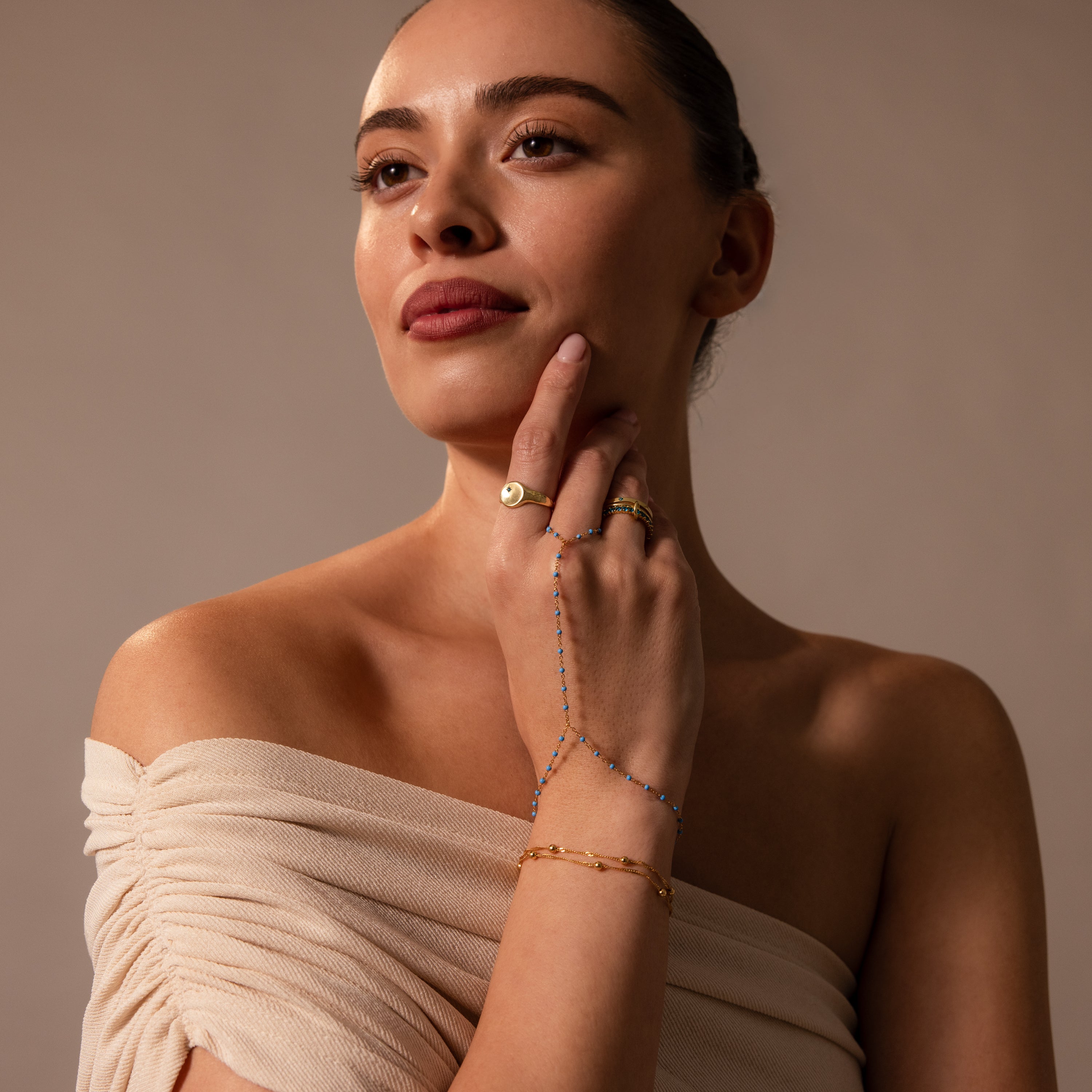 Woman in an off-the-shoulder beige top wears gold jewelry and the Ocean Beaded Hand Chain Bracelet, gazing thoughtfully to the side.