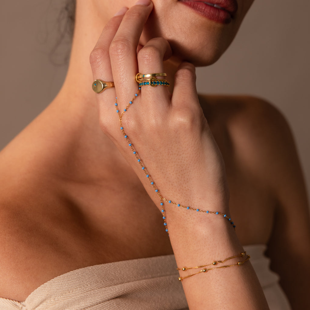 A woman wearing gold rings and the Ocean Beaded Hand Chain Bracelet in 18K Gold touches her face against a neutral background.