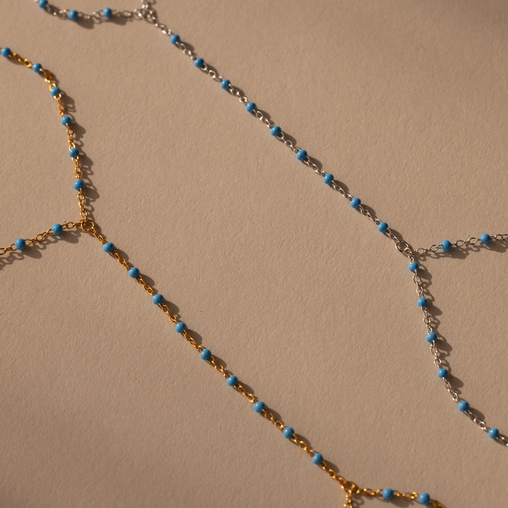 The Ocean Beaded Hand Chain Bracelet features gold and silver delicate chains with small turquoise beads, beautifully arranged on a beige surface to evoke serene ocean vibes.