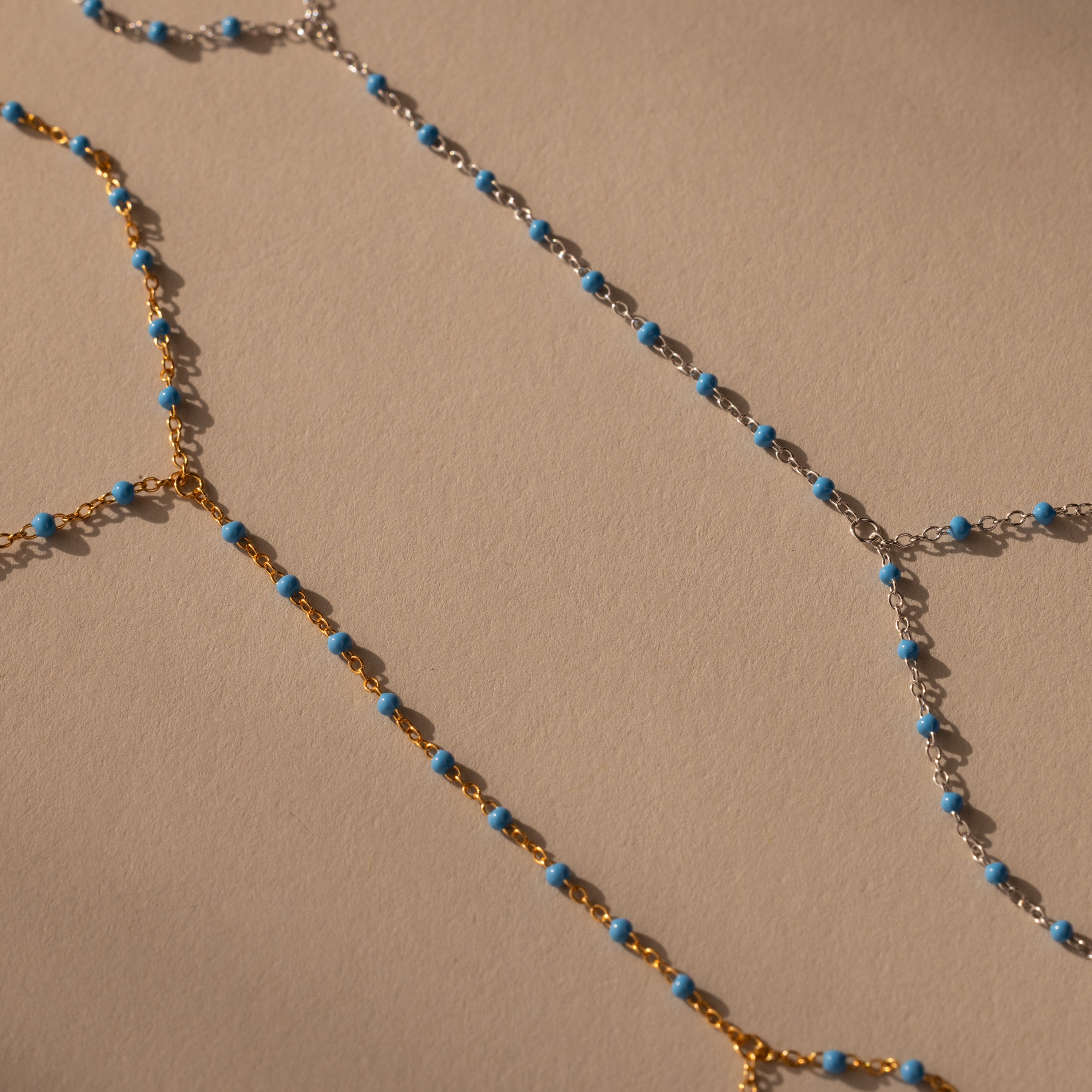 The Ocean Beaded Hand Chain Bracelet features gold and silver delicate chains with small turquoise beads, beautifully arranged on a beige surface to evoke serene ocean vibes.