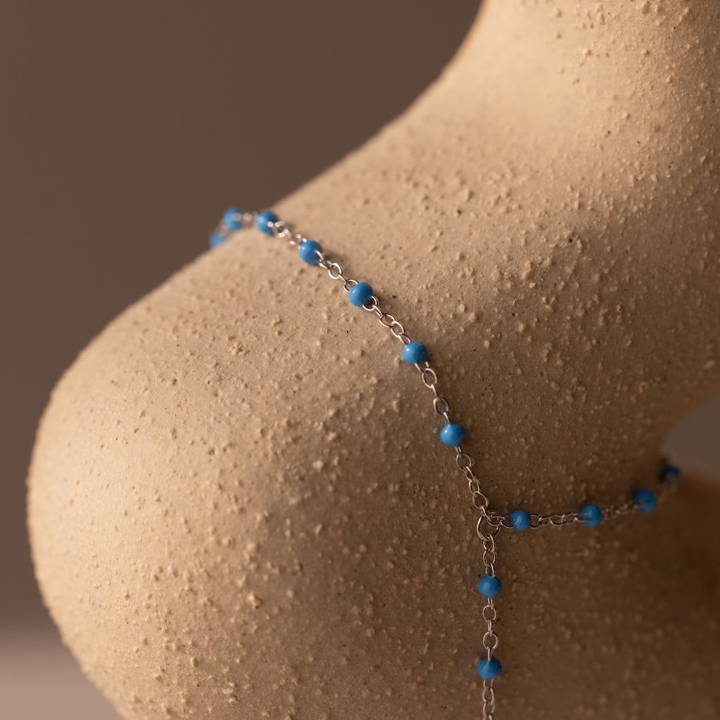 The Ocean Beaded Hand Chain Bracelet, featuring turquoise beads on a delicate chain, drapes elegantly over a curved, textured beige sculpture.