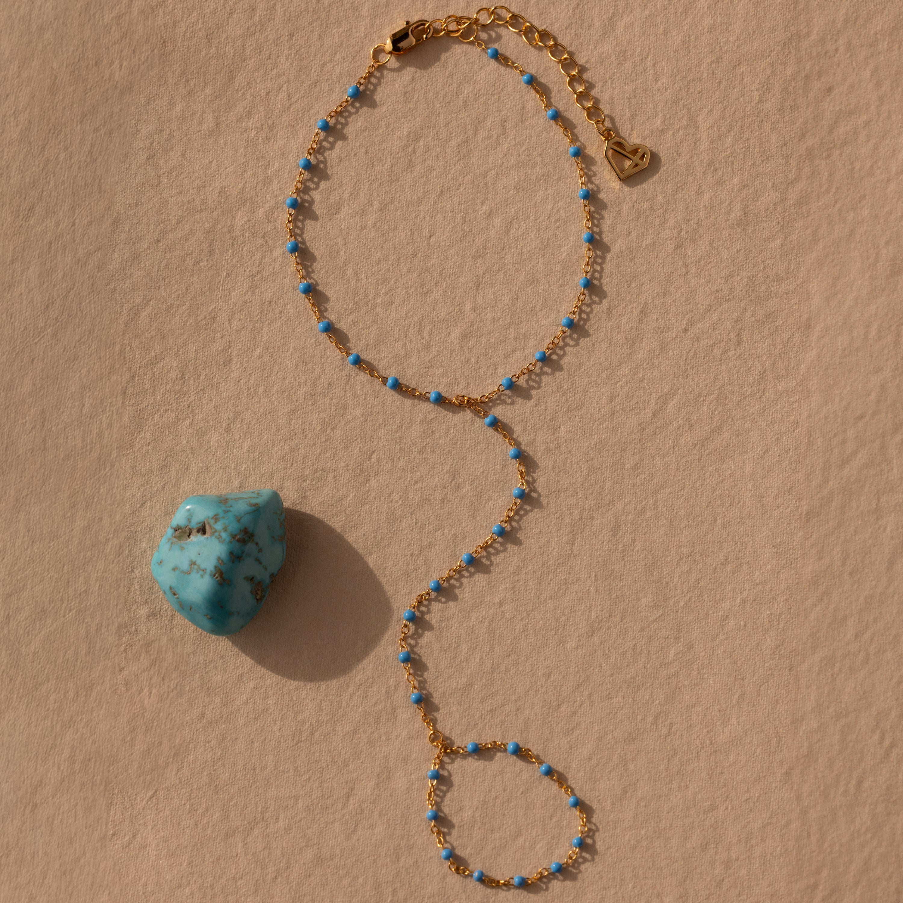 The Ocean Beaded Hand Chain Bracelet in 18K Gold, adorned with small blue beads, is displayed next to a turquoise stone on a beige textured surface.