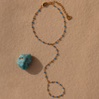 The Ocean Beaded Hand Chain Bracelet, a gold delicate chain with small blue beads, rests by turquoise on a beige textured surface, capturing the charm of ocean-inspired jewelry.