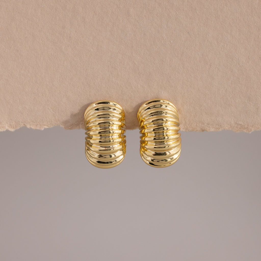 A pair of chunky, gold-toned ribbed hoop earrings displayed against a minimalist backdrop. The hoops feature a bold, domed silhouette with a high-polish finish and evenly spaced horizontal ridges that wrap around the surface, creating a sculptural, pillowy texture