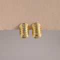 A pair of chunky, gold-toned ribbed hoop earrings displayed against a minimalist backdrop. The hoops feature a bold, domed silhouette with a high-polish finish and evenly spaced horizontal ridges that wrap around the surface, creating a sculptural, pillowy texture